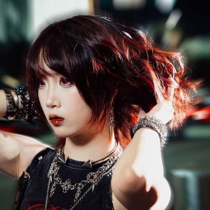 A woman posing in a black graphic tank top with red lettering, accessorized with silver chains and bracelets, set against a blurred city street background with motion blur.